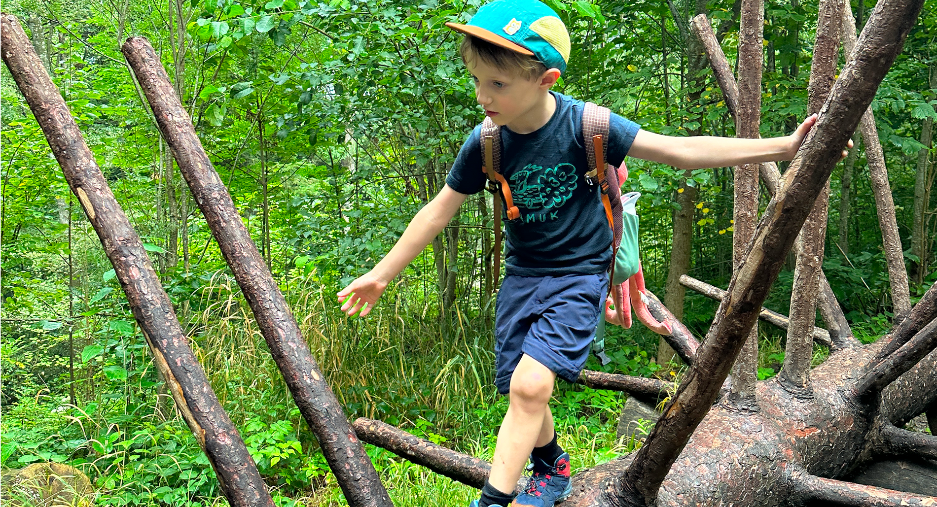 Junge in Wanderkleidung klettert im Grünen über einen Baumstamm
